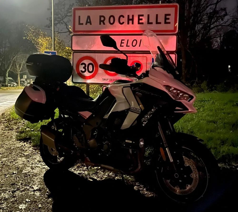 Une moto Kawasaki Versys 1100 SE blanche avec des valises et un top-case garée devant des panneaux d'informations routières et le panneau d'entrée de la ville La Rochelle.