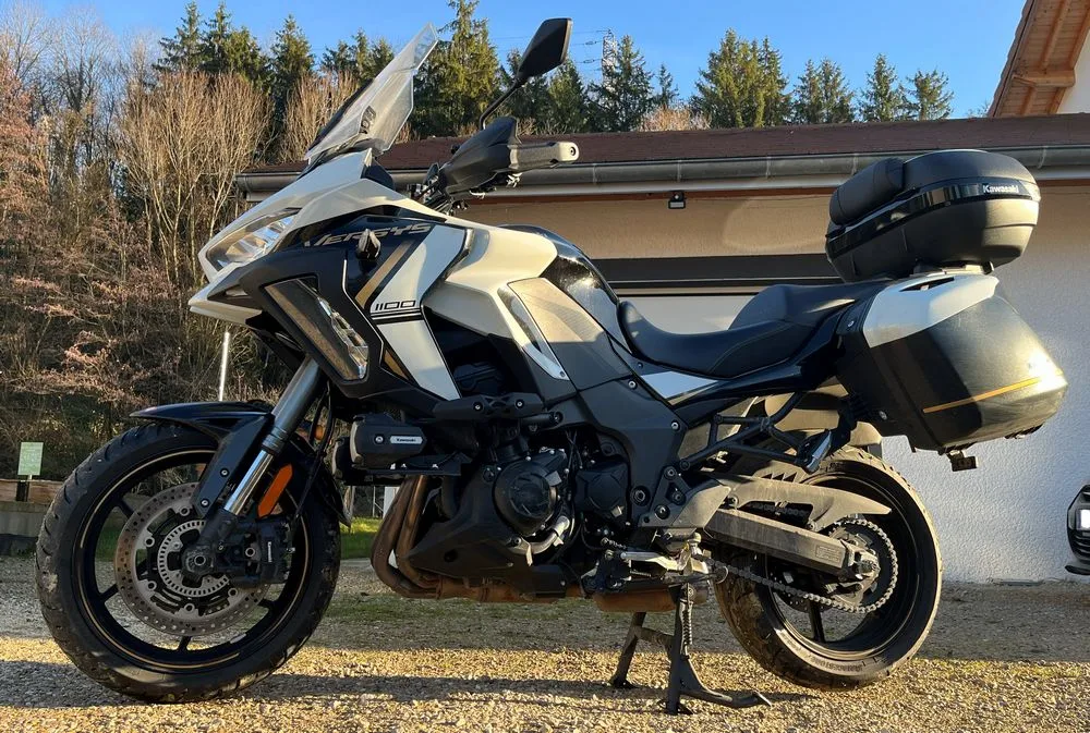 Une moto Kawasaki Versys 1100 SE blanche avec des valises et un top-case garée devant une maison avec du gravier et une foret
