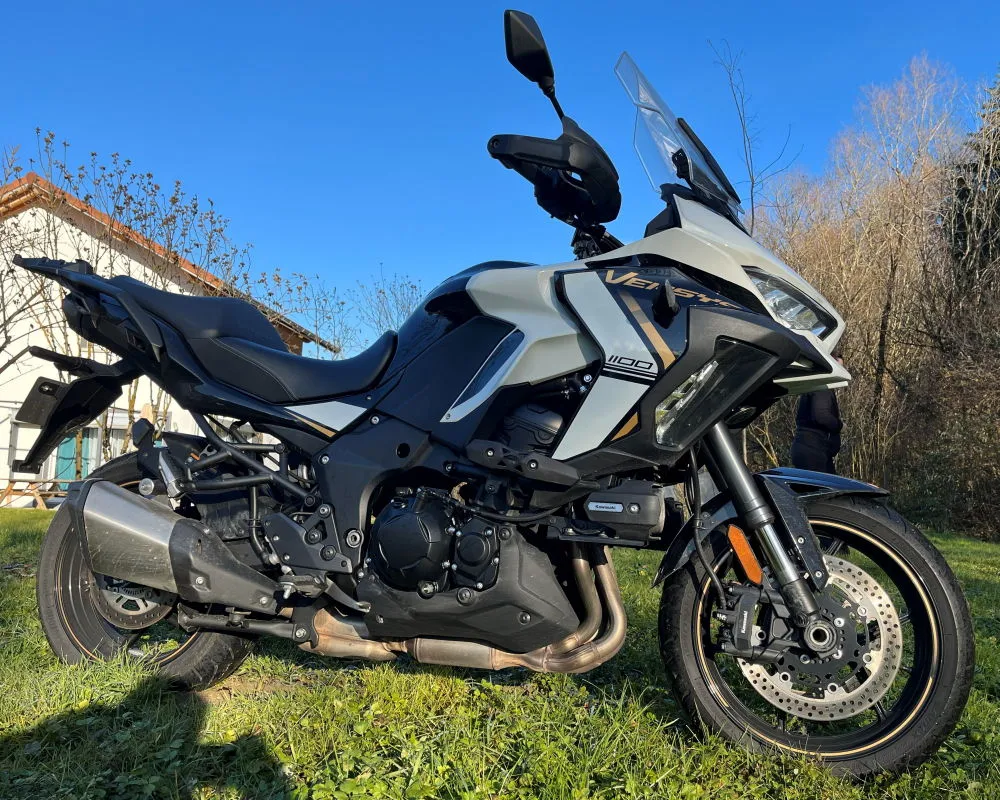 Une moto Kawasaki Versys 1100 SE blanche avec des valises et un top-case garée dans un jardin, de l'herbe verte, des arbustes et un ciel bleu