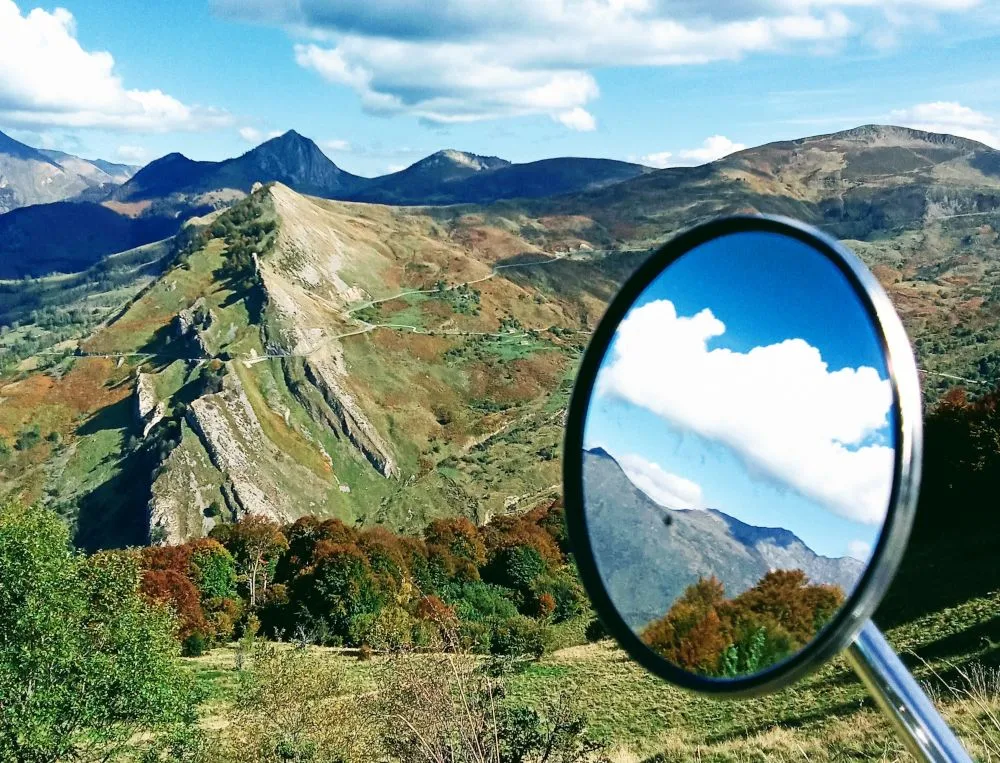 Un rétroviseur rond de moto, une vallée de montagne, des nuages et un ciel bleu.