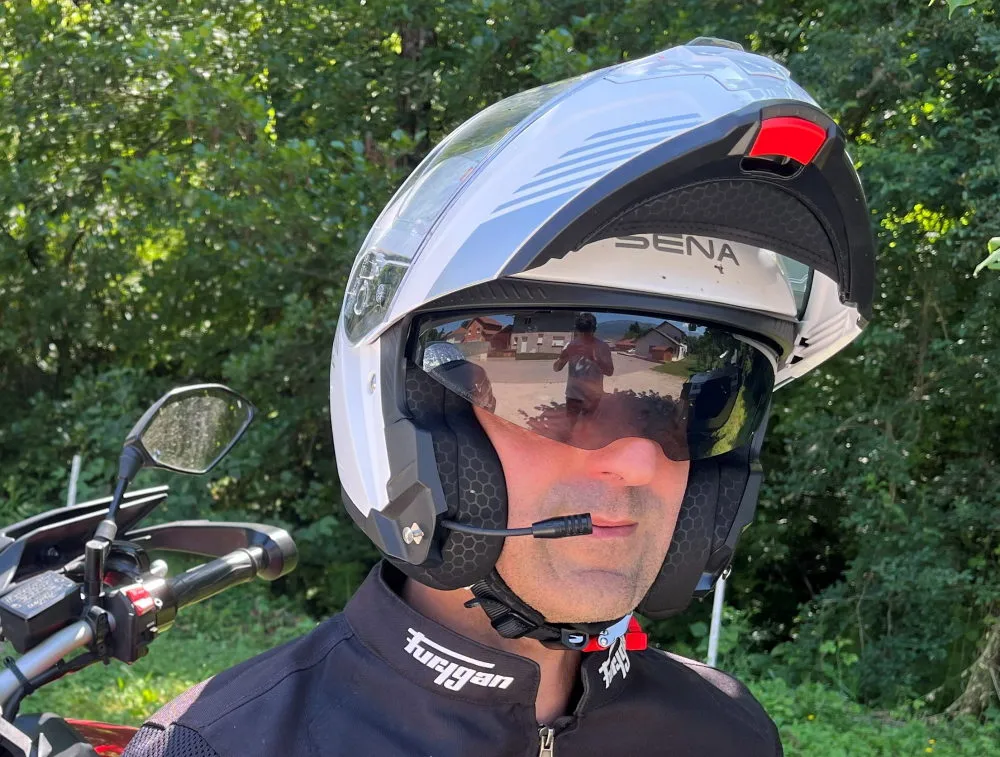 Un casque moto modulable Sena Specter Blanc en position ouverte avec l'écran solaire, de la végétation