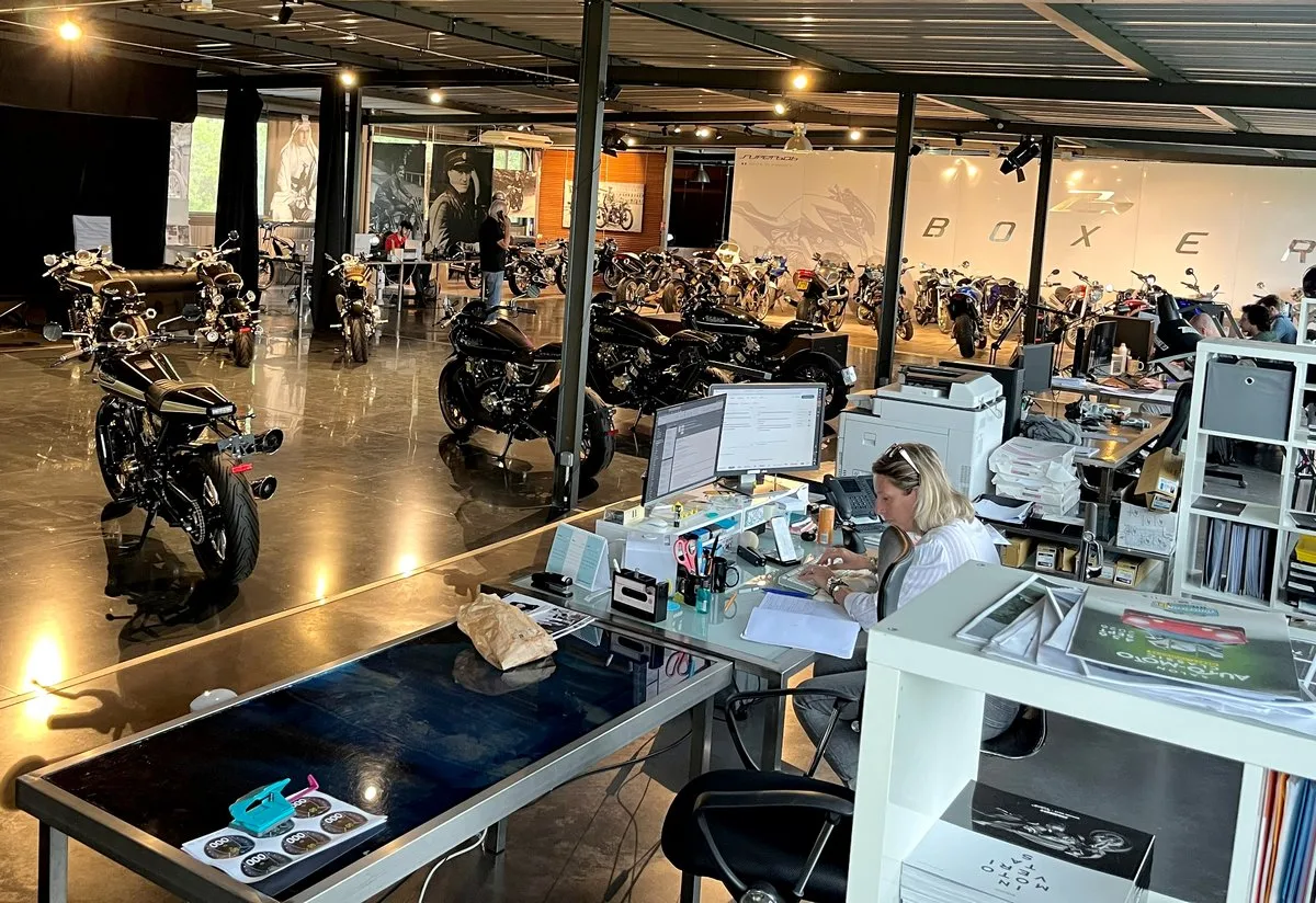 Un open space, plusieurs motos de prestige brough superior, un bureau, une secrétaire blonde