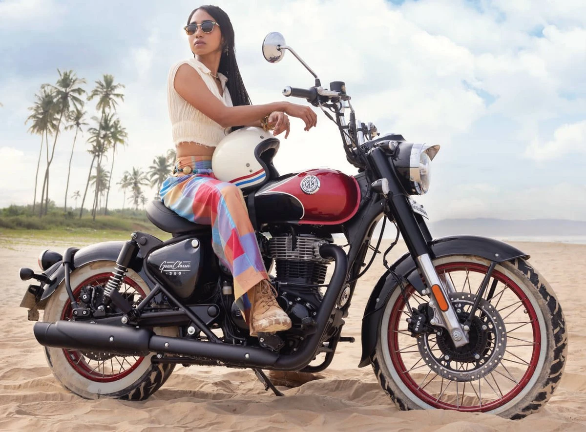 Une moto Royal-Enfield Goan Classic 350 rouge et noir, une plage, des palmiers, une jeune fille brune avec un pantalon multicolore et un top blanc