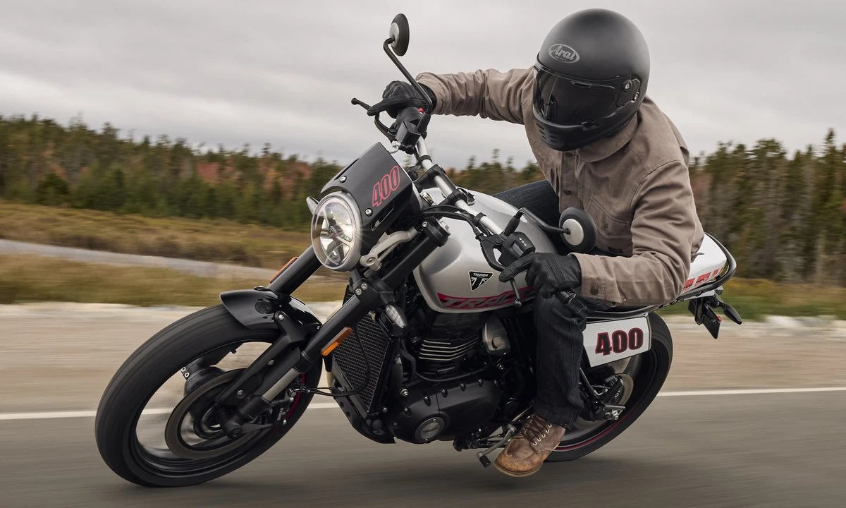 Une Triumph 400 Tracker sur une route, un motard avec un casque noir et une foret derrière.