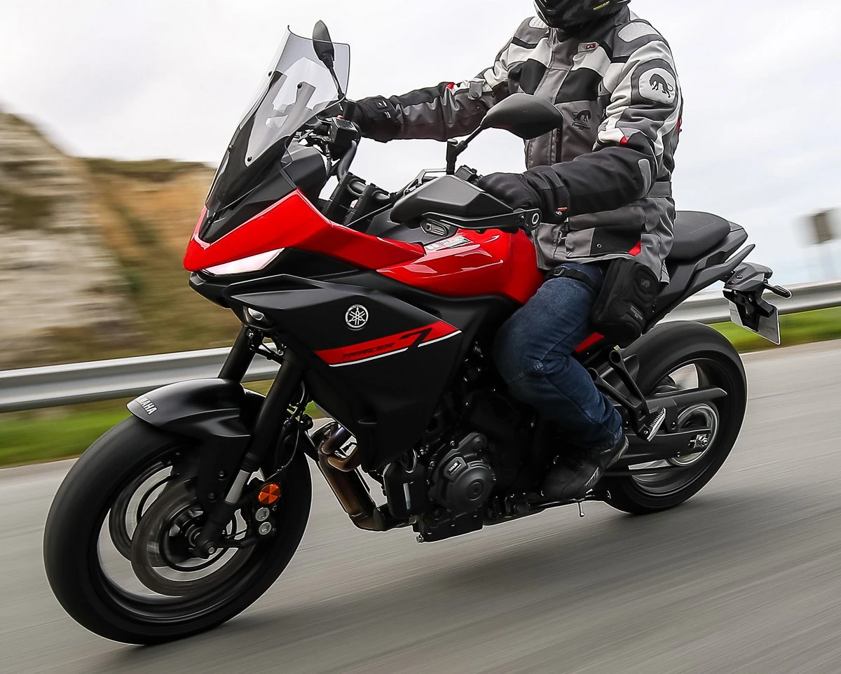 Une moto Yamaha Tracer 7 rouge et noire, une route, une barrière de sécurité, des herbes hautes, un fond de mer et un pilote habillé en vêtements gris